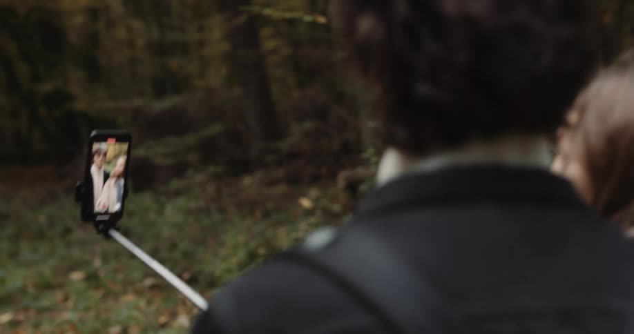 A young couple are hiking through the woods holding their phone out in front of them on a selfie stick