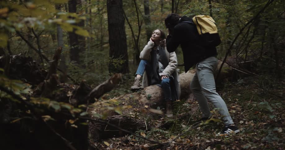 A man uses a camera to photograph a woman as she poses on a fallen tree trunk in a forest during the day