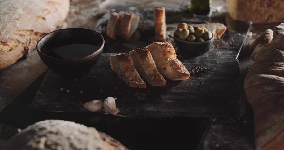 A wooden slab has slices of bread olives and a few other food items on it and is surrounded by loaves of bread