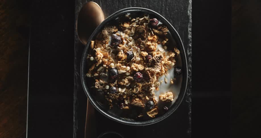 A still shot of granola and fruit with milk in a bowl with a bronze spoon next to the bowl