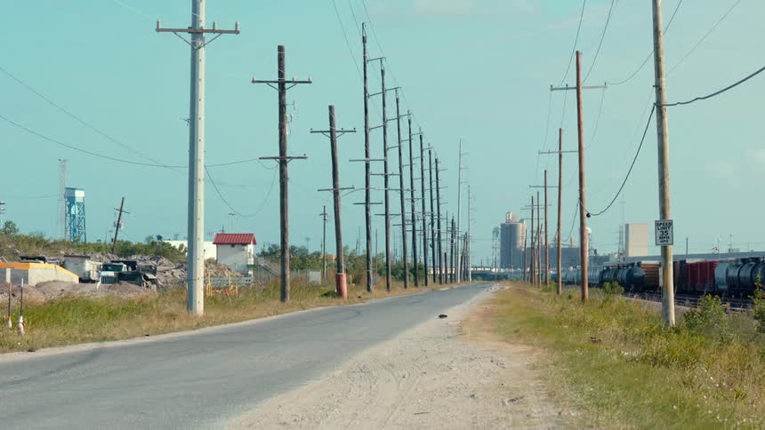A road lined with electrical poles near a train and factory