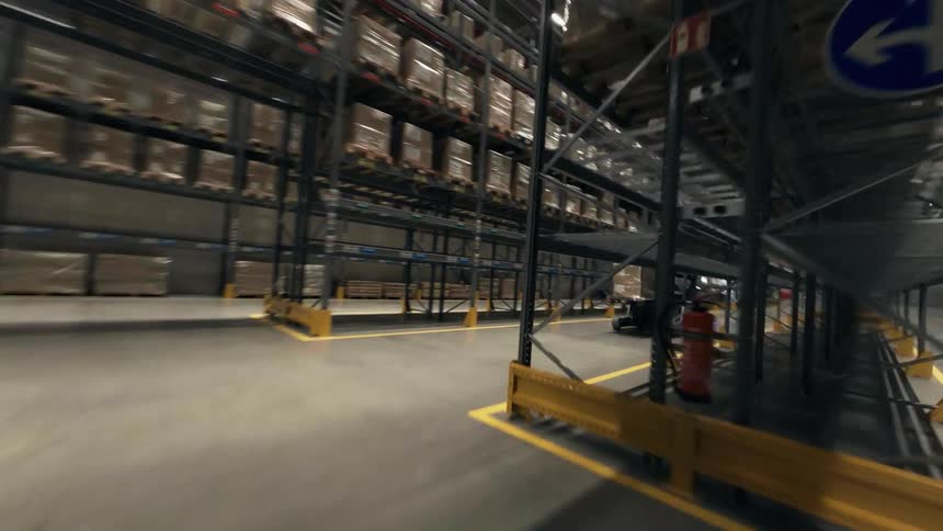 An FPV drone shot zooming through a large warehouse, weaving through metal racks, and flying above a worker driving a forklift