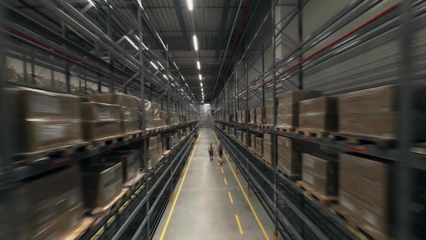 An FPV drone shot moving down the aisles of a large warehouse that passes over two workers moving a box