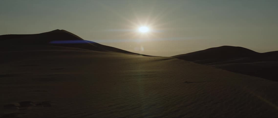 The sun shines brightly in a hazy sky over desert dunes and mountains