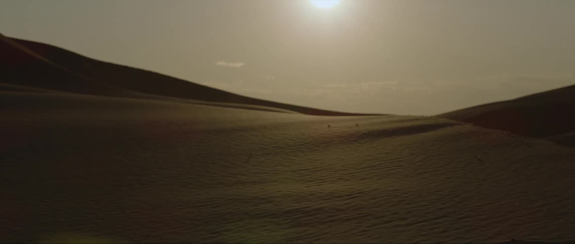 A wide open aerial view of the desert is shown with sandy hills and the sun shining