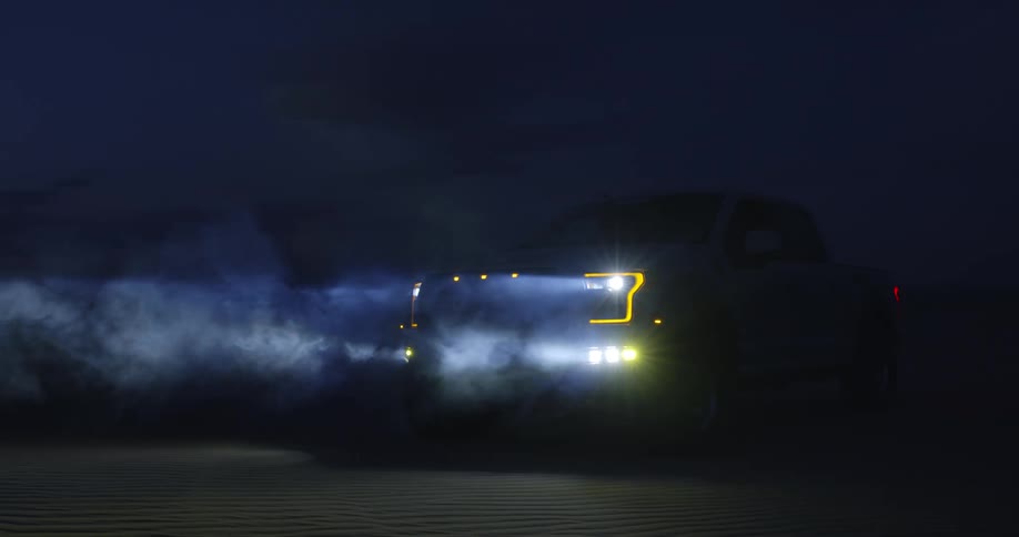 A truck is parked in the darkness and the headlights come on and smoke moves in the light beams