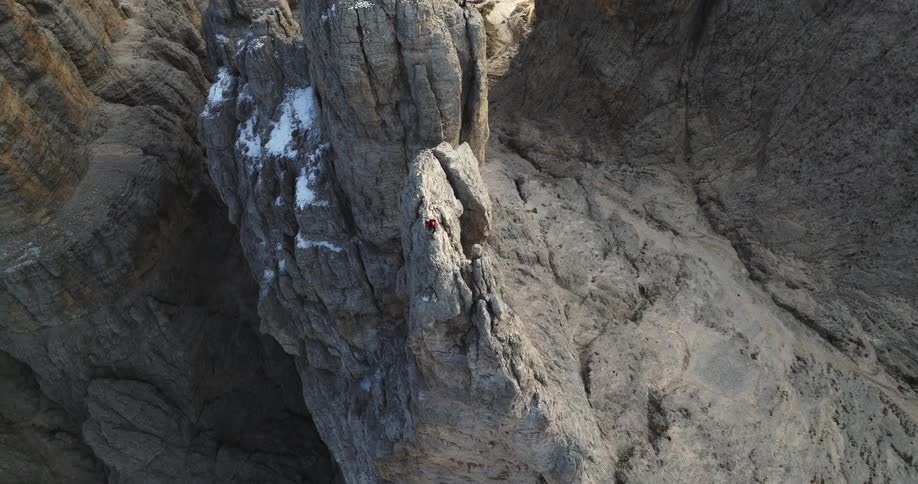 Snow dusts cliff near climber with building on barren plateau nearby
