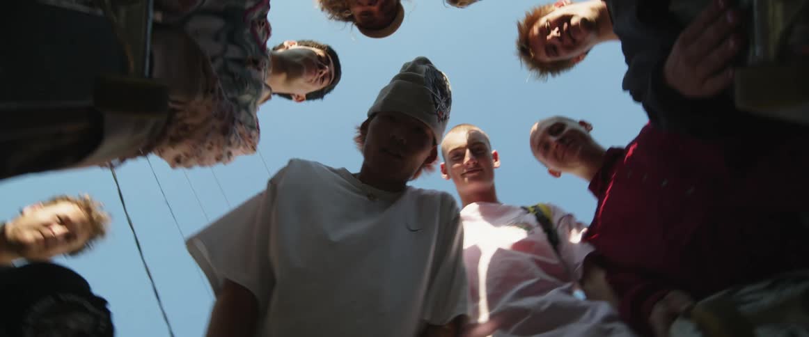 A small group of teenage boys stand in a circle with skateboards in hand as they are filmed going in circles from the bottom up