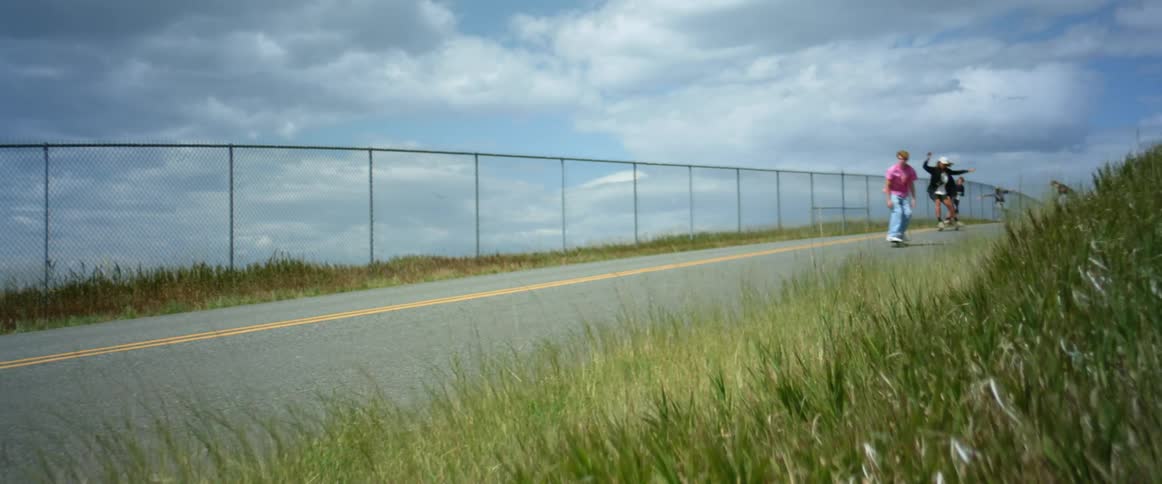 A group of teenage boys are riding their skateboards down an open road along a chain link fence under a cloudy sky