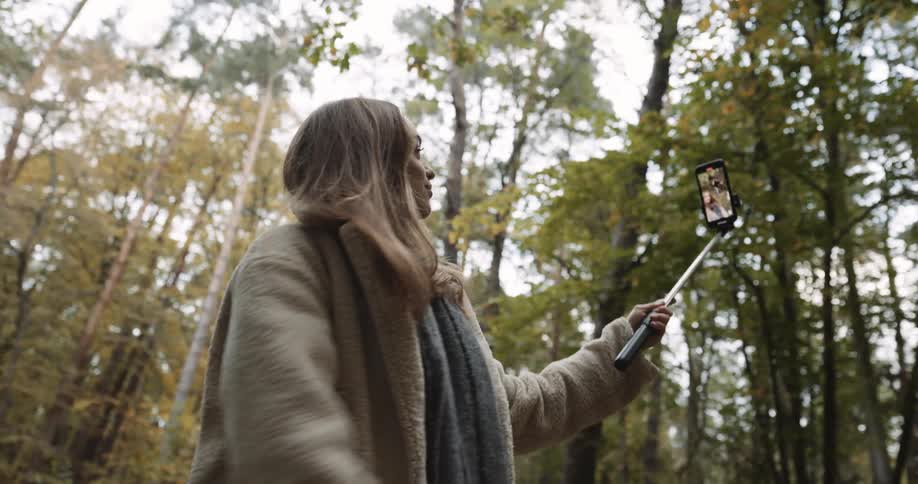 A teenage couple are walking on a trail through the woods and she is holding her phone on a selfie stick and she stops to pose for the camera