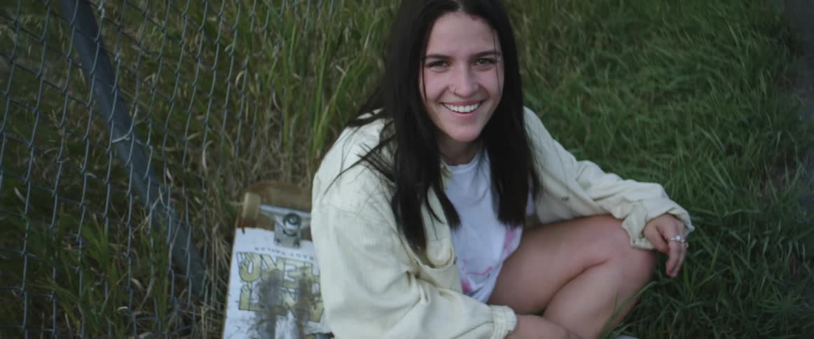A smiling woman sitting on the grass with a skateboard at her side
