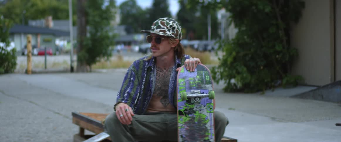 Man holding painted skateboard sits on bench behind building