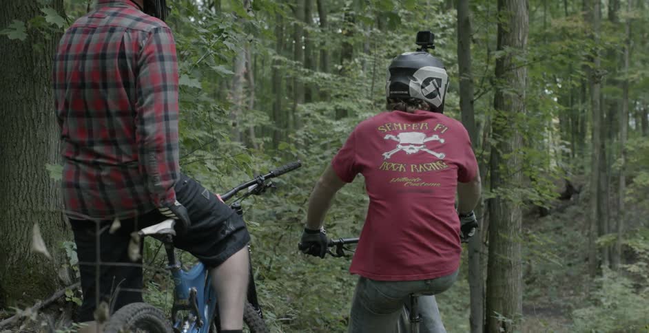 Two men sit on their bikes before a forest