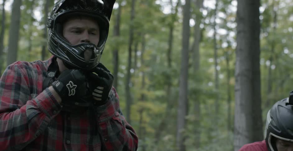 Two men put on helmets and goggles while in the woods during the day