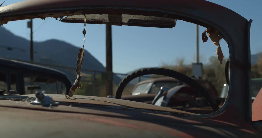 Rusted car sits in junkyard as vehicle in background passes between nearby mountain and junkyard