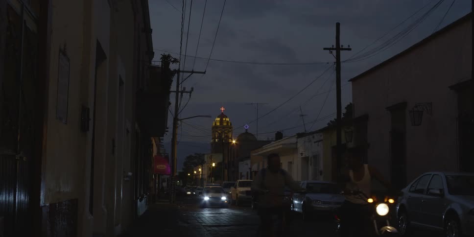 A dark side street of a town with parked cars and minor traffic at dusk