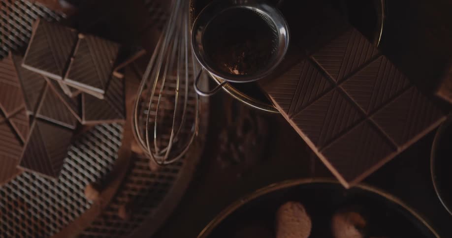 Chocolate candy is in a bowl on a table next to square chocolate cookies and a wire whisk
