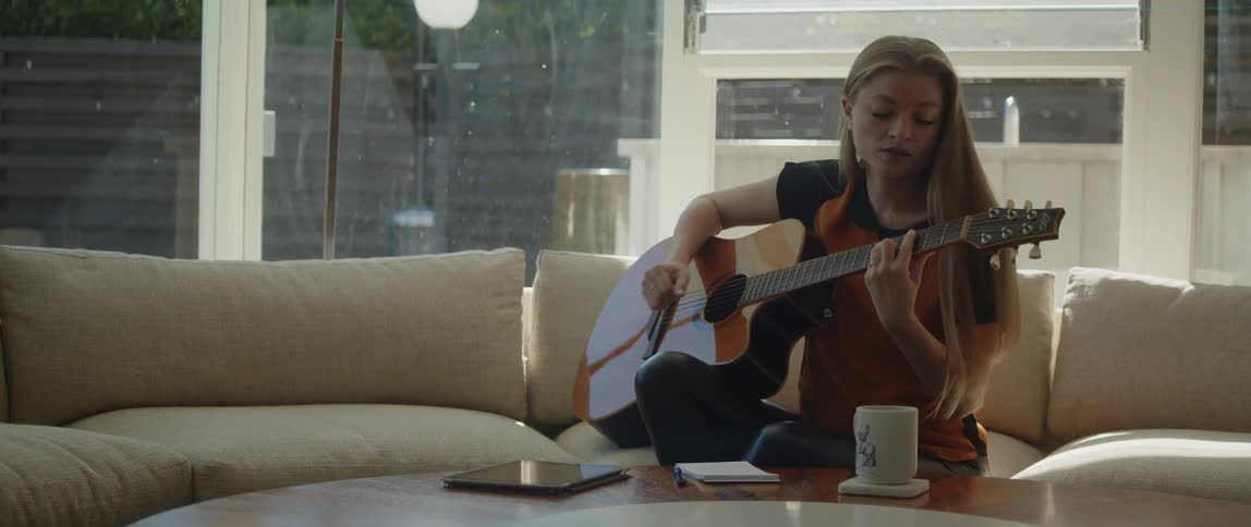 A blonde woman playing the guitar sitting on a beige couch while singing