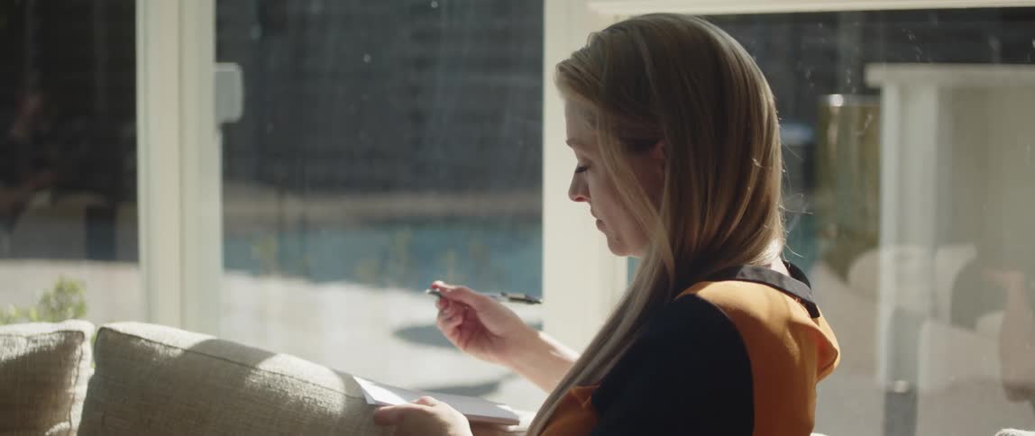 A woman sitting on a sofa thinks for a moment before beginning to write on a notepad