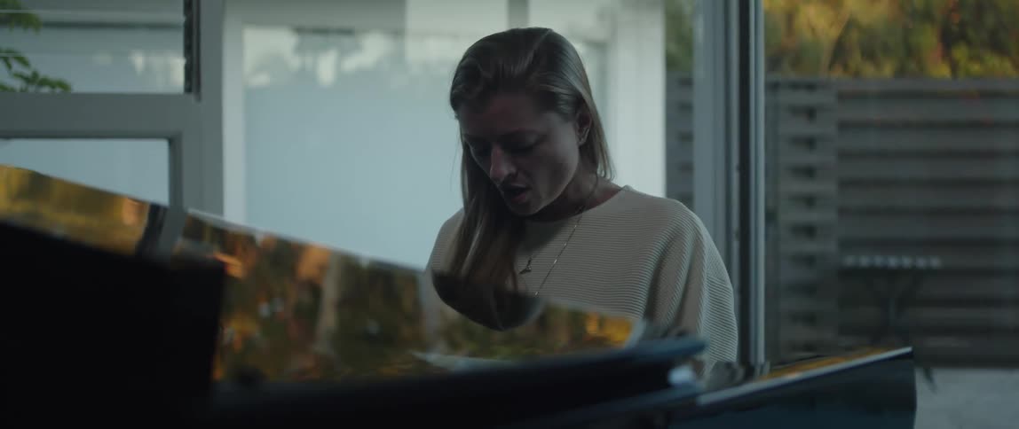 Blonde woman wearing a white sweater and necklace plays grand piano in room singing