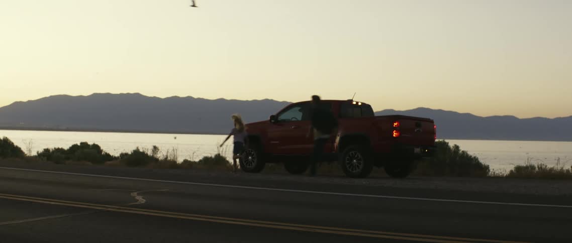 A man chases after a little girl running around a truck with water and mountains in the background