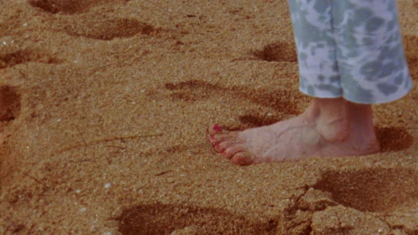 Close up of the feet of a barefoot person walking on the sand of a beach caught on vintage film