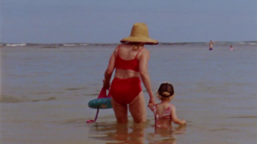 Mother in red bikini and hat holds daughters hand and walks into the sea