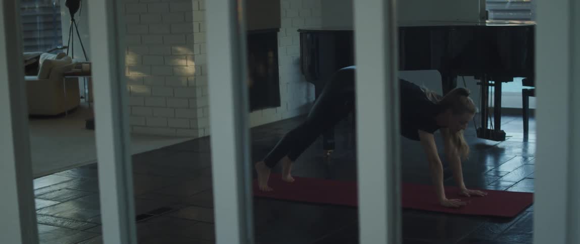 Blonde woman with ponytail and in black shirt and yoga pants does yoga on yoga pants next to grand piano