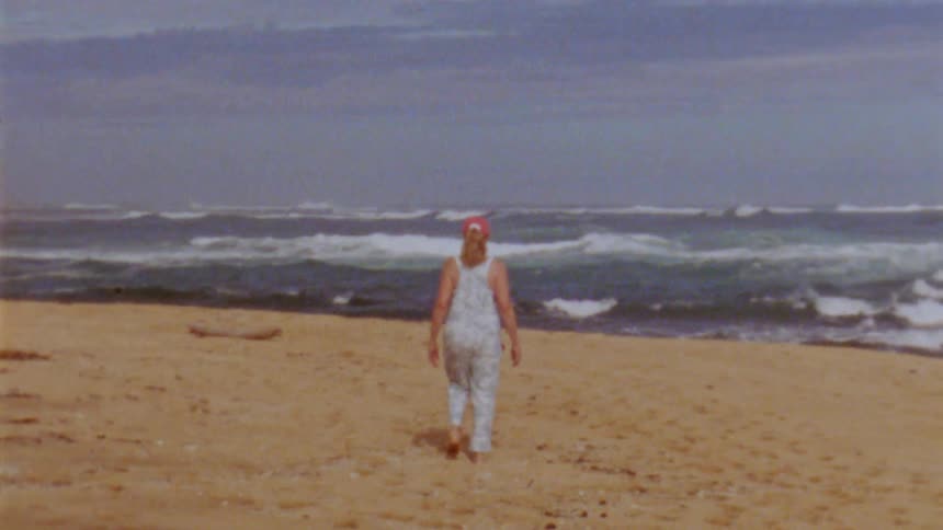 Young woman wearing overalls walking over the sand beach next to the ocean caught on vintage film