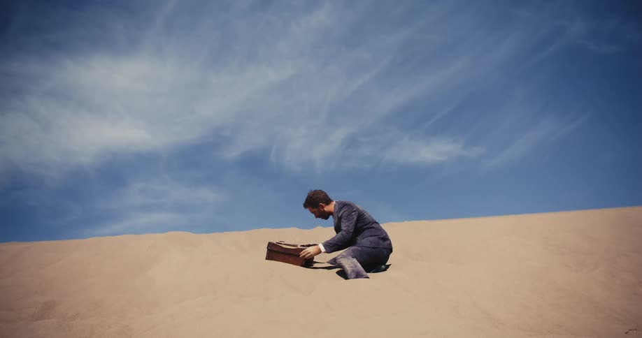 A man is sitting in the sand on a desert hill as he opens a briefcase
