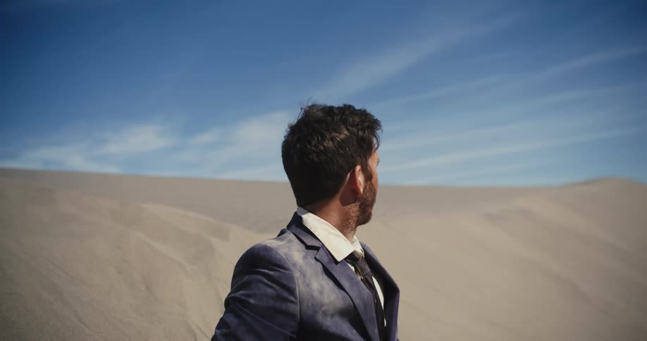 The sun is shining on the face of a man in a dirty suit standing in the desert looking around tired and confused