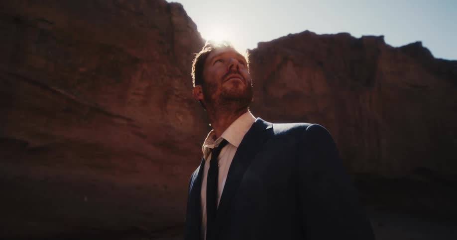 A man in a suit looks around at his surroundings while standing in a canyon on a sunny day