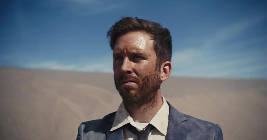 The sun is shining on the face of a man in a dirty suit standing in the desert looking tired and confused