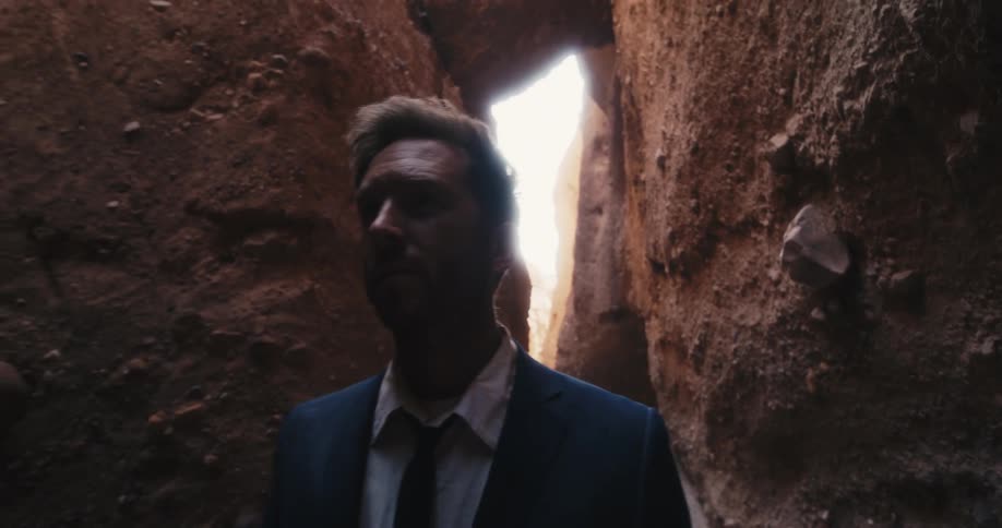 Man wearing suit walks along narrow gap between cliff walls near arch and overhang
