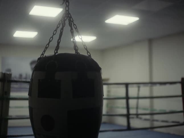 Black punching bag swinging gently from ceiling near a ring in the gym
