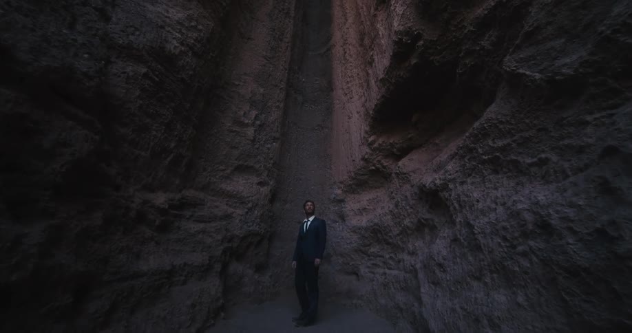 A man in a suit is standing inside a desert mountain crevasse as light shines from above