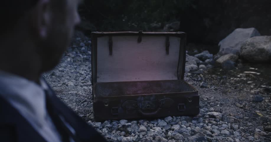 A man in a suit sitting in the gravel beside a river slowly turns his head to look at an open suitcase sitting nearby