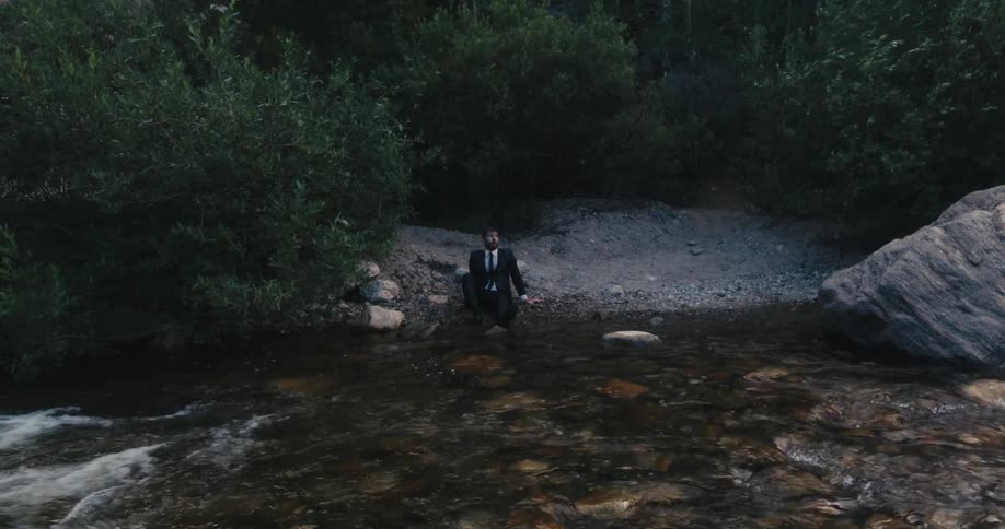 Man in business suit sits on ground at edge of quickly flowing river breathing deeply