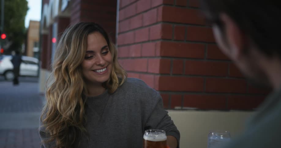 Man wearing green shirt and ear tunnel plugs clinks glasses with woman with brown hair and gray sweater sitting at table in front of cafe
