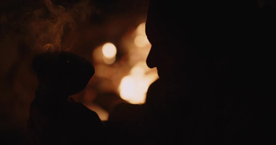 Man silhouetted against campfire holds smoking bundle up and bows his head