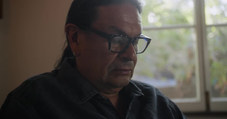 Man with long hair earrings glasses and black shirt sits in armchair reading a book in room