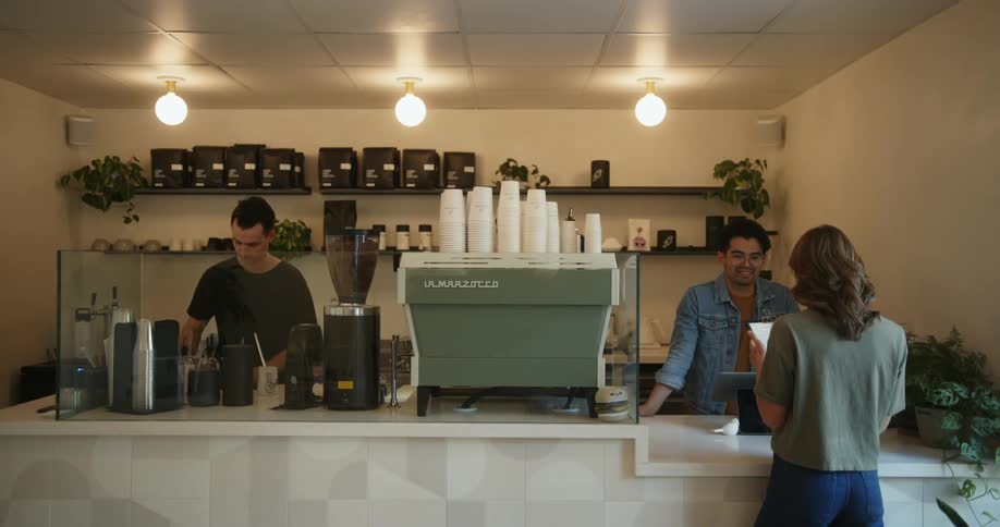 Woman with brown hair green t shirt and jeans enters cafe and heads to cafe bar to look at menu next to baristas working in cafe