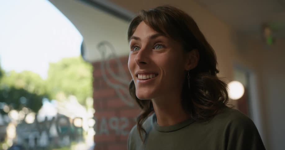 Woman with brown wavy hair green eyes earrings and green t shirt reads menu and talks to person smiling in front of cafe