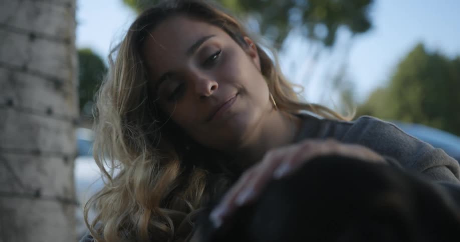 Woman with brown wavy hair and green eyes strokes black dog in the street
