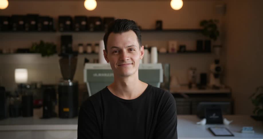 Barista with black t shirt and wristwatch looks at camera in front of cafe bar