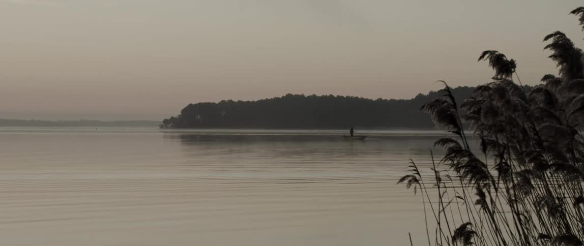 A fisherman is standing in a small boat in a lake surrounded by forests under a hazy sky