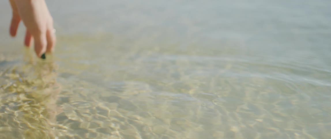 Sunlight reflects on ripples in the water as a woman with painted fingernails and wearing rings and a bracelet is moving her hand across the water