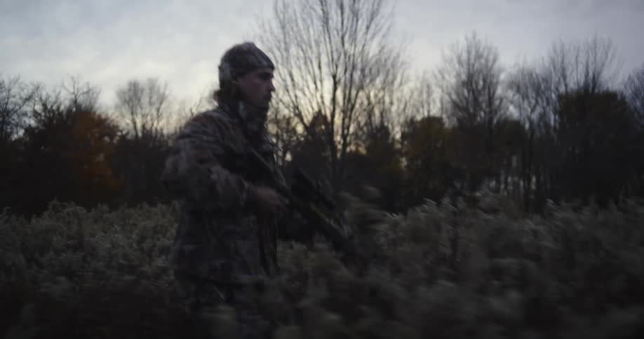 A man walks through a landscape filled with bushes and aims a weapon