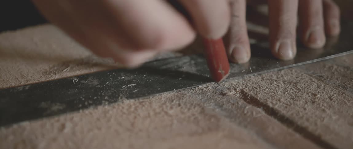 Someone is holding down a ruler as they mark a line on a piece of wood covered in sawdust