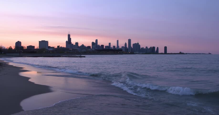 Waves roll over the sand on a beach as tall buildings on the city skyline are in the distance under a clear sky